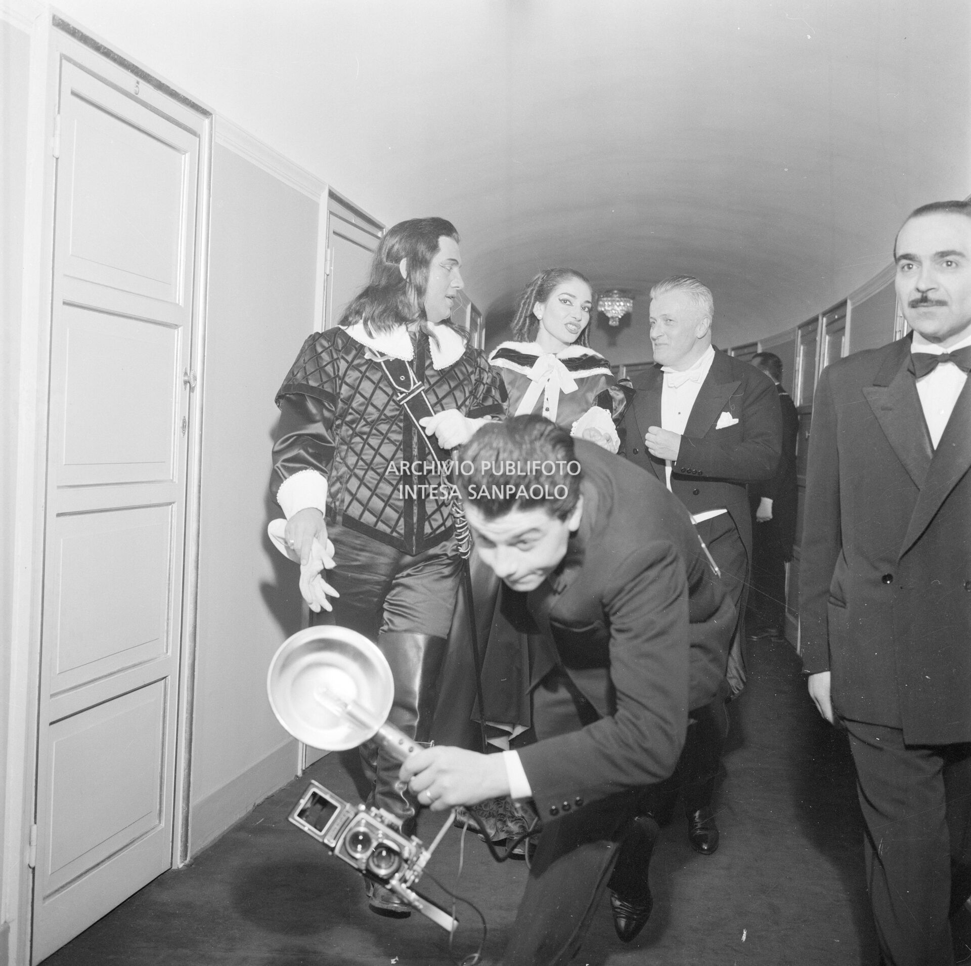 Un fotoreporter con macchina fotografica precede di corsa la soprano Maria Callas, il baritono Ettore Bastianini, in costume di scena, e il direttore d'orchestra Gianandrea Gavazzeni al Teatro alla Scala la  sera dell'inaugurazione della stagione lirica 1957-1958 con l'opera "Un ballo in maschera", di Giuseppe Verdi, diretta dallo stesso Gavazzeni, con la regia di Margherita Wallmann;